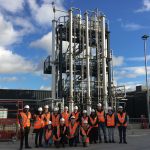 a group of students wearing orange hi vis vests and hard hats stand near a large metal Amine capture plant